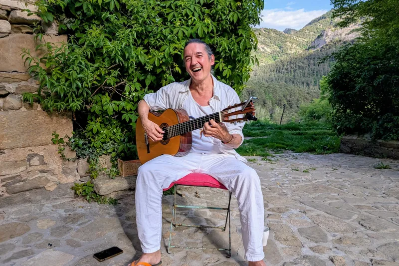 Manu riendo con la guitarra en un patio de piedra, entre montañas